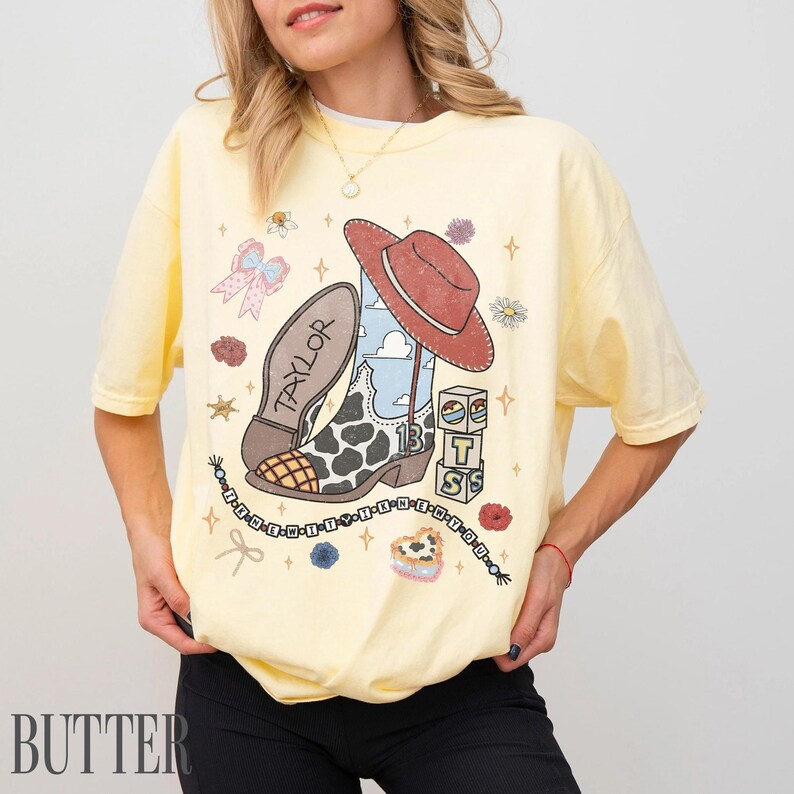 May include: A pale yellow short-sleeve t-shirt featuring a graphic print of a cowboy boot with cow print, a red cowboy hat, blocks with the letters T, S, and a number 13, a bow, and a beaded string with the text IT-THE-NEW-IT-THE-NEW-YOU.