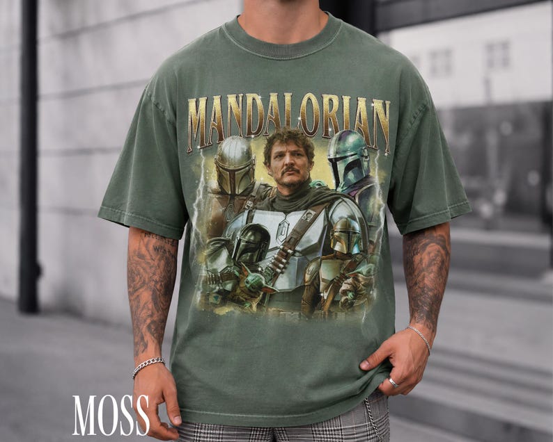 May include: Moss green t-shirt with a graphic of characters from the Mandalorian series. The word "MANDALORIAN" is printed across the top in gold lettering. The shirt has a vintage style and a distressed appearance.