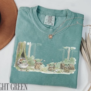Pu&ograve; includere: T-shirt verde chiaro con una grafica di Grogu da The Mandalorian. Il design presenta Grogu in varie pose in un ambiente forestale. La maglietta ha un girocollo e maniche corte. Una collana d'oro &egrave; drappeggiata sulla maglietta.