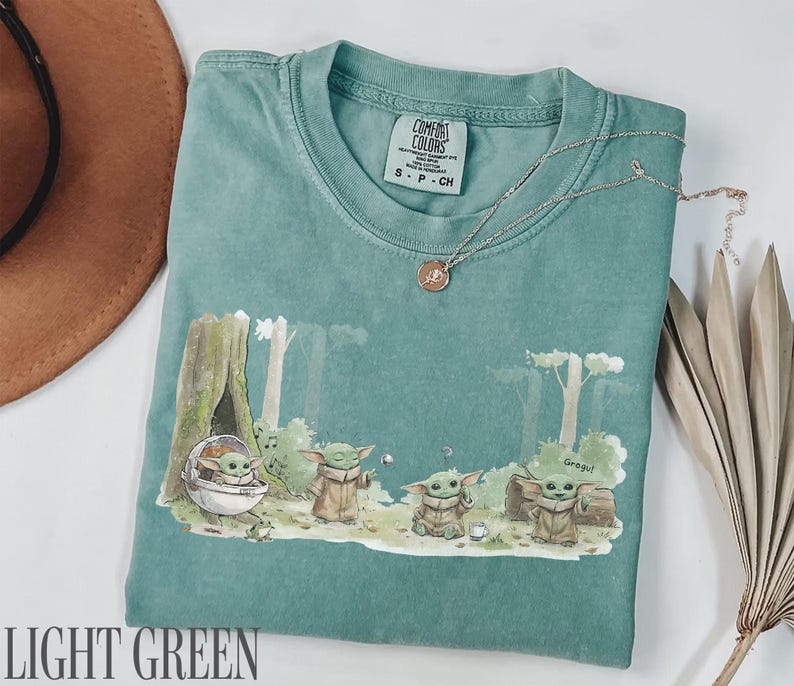 Pu&ograve; includere: T-shirt verde chiaro con una grafica di Grogu da The Mandalorian. Il design presenta Grogu in varie pose in un ambiente forestale. La maglietta ha un girocollo e maniche corte. Una collana d'oro &egrave; drappeggiata sulla maglietta.
