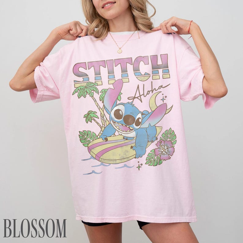 May include: A light pink t-shirt featuring a graphic of Stitch surfing on a surfboard with the words "STITCH Aloha". The design includes palm trees, waves, and flowers. The word "BLOSSOM" is printed at the bottom.