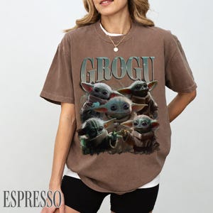K&ouml;nnte beinhalten: Espressofarbenes T-Shirt mit einer Grafik von Grogu, auch bekannt als Baby Yoda, mit dem Wort "GROGU" &uuml;ber dem Bild. Das Design enth&auml;lt mehrere Bilder von Grogu. Das T-Shirt wird von einer Person getragen.