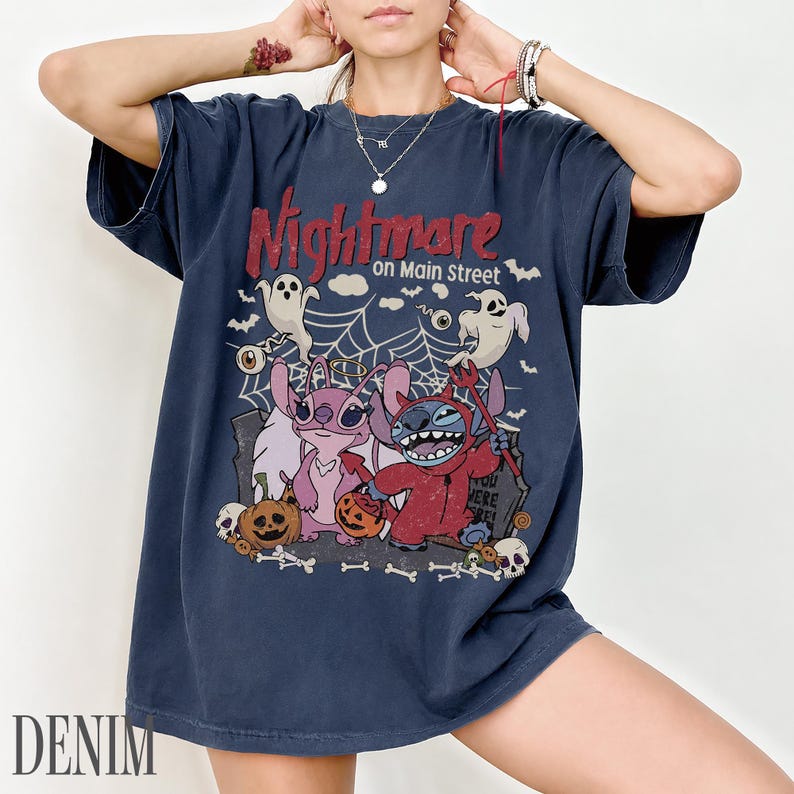 May include: A dark blue short-sleeve t-shirt featuring a graphic print with the text Nightmare on Main Street. The design includes two cartoon characters, one dressed as a devil and one with a halo, surrounded by ghosts, spiderwebs, and pumpkins.