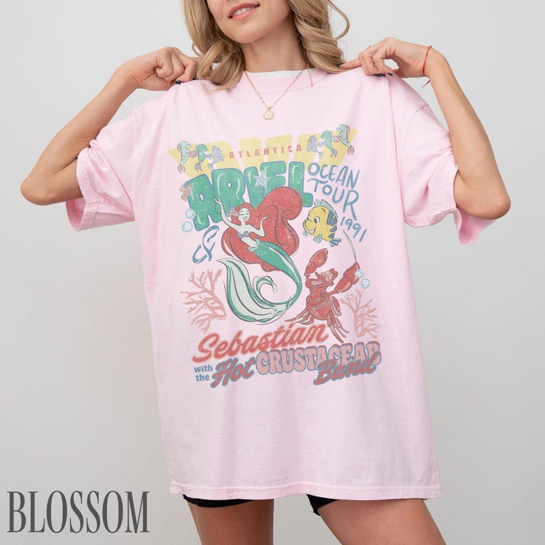 Puede incluir: Una camiseta de manga corta de color rosa claro con un estampado gr&aacute;fico de Ariel, Flounder y Sebasti&aacute;n. El texto dice Atlantica Ocean Tour 1991, Ariel, Sebastian with the Hot Crustacean Band. El dise&ntilde;o incluye ilustraciones de coral y burbujas.