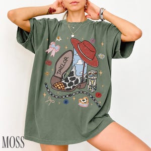 May include: An olive green short-sleeve t-shirt featuring a graphic print of a cow-print cowboy boot, a red hat, a pink bow, and stacked blocks with the letters T, S, and the number 13. The text TAYLOR is on the boot sole.