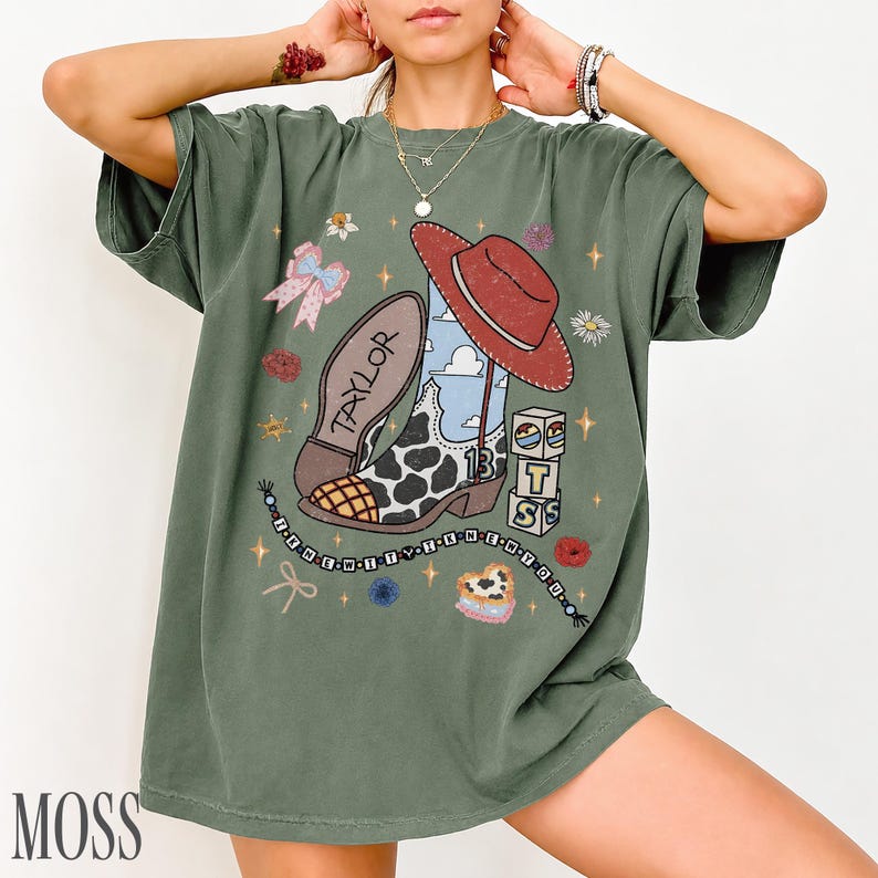 May include: An olive green short-sleeve t-shirt featuring a graphic print of a cow-print cowboy boot, a red hat, a pink bow, and stacked blocks with the letters T, S, and the number 13. The text TAYLOR is on the boot sole.