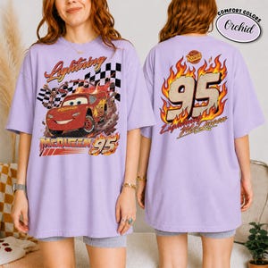 May include: Lavender t-shirt featuring Lightning McQueen from the movie Cars. The front graphic shows McQueen racing with the text "Lightning McQueen 95". The back of the shirt has "95" in flames and "Lightning McQueen" text.