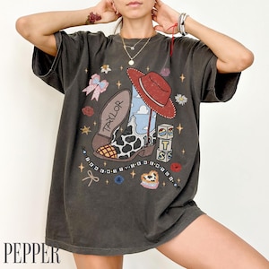 May include: A charcoal gray short-sleeve t-shirt featuring a graphic print of a cow-print cowboy boot with a red hat, the name Taylor, the number 13, stacked blocks with the letters T, S, and a beaded necklace design with the text I KNEW YOU WERE TROUBLE.