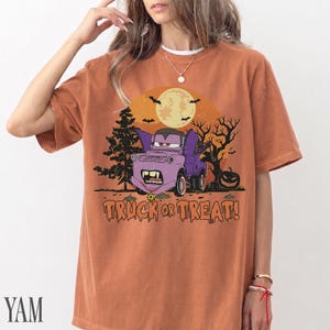 Może przedstawiać: Pomarańczowa koszulka z kr&oacute;tkim rękawem z grafiką o tematyce Halloween. Grafika przedstawia fioletowy samoch&oacute;d z kresk&oacute;wek z wampirzymi kłami, pełnię księżyca, nietoperze i napis "TRUCK OR TREAT!".