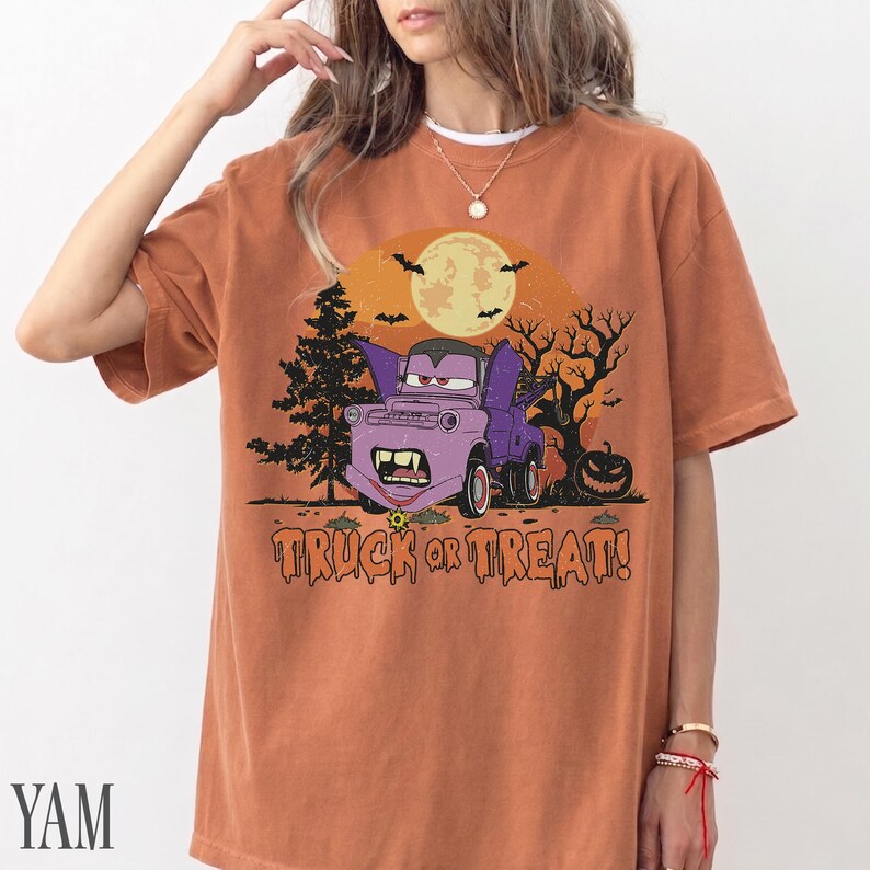 Może przedstawiać: Pomarańczowa koszulka z kr&oacute;tkim rękawem z grafiką o tematyce Halloween. Grafika przedstawia fioletowy samoch&oacute;d z kresk&oacute;wek z wampirzymi kłami, pełnię księżyca, nietoperze i napis "TRUCK OR TREAT!".