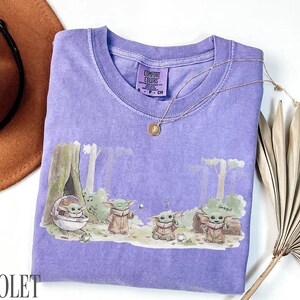 Pu&ograve; includere: Maglietta Comfort Colors viola con una scena della foresta con personaggi di Grogu. La maglietta ha un girocollo e maniche corte. Una collana d'oro con un ciondolo &egrave; sulla maglietta. Un cappello marrone e una pianta secca sono sullo sfondo.