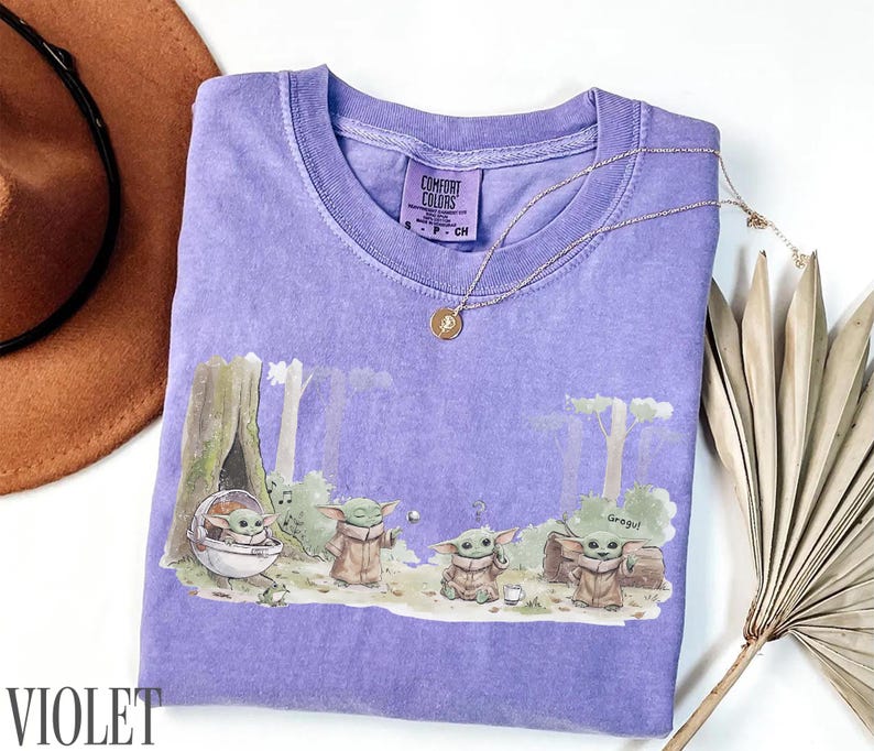 Pu&ograve; includere: Maglietta Comfort Colors viola con una scena della foresta con personaggi di Grogu. La maglietta ha un girocollo e maniche corte. Una collana d'oro con un ciondolo &egrave; sulla maglietta. Un cappello marrone e una pianta secca sono sullo sfondo.