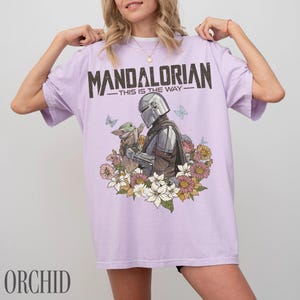 May include: Lilac t-shirt with a graphic of The Mandalorian and Grogu, framed by flowers and butterflies. The text "MANDALORIAN - THIS IS THE WAY" is printed across the top. A comfortable and stylish tee.
