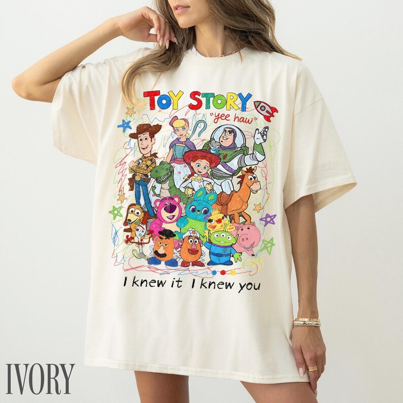 May include: An ivory t-shirt featuring a colourful graphic print of various Toy Story characters including Woody, Buzz Lightyear, and Jessie. The design includes the text "TOY STORY", "yee haw", and "I knew it I knew you" surrounded by scribbled stars.