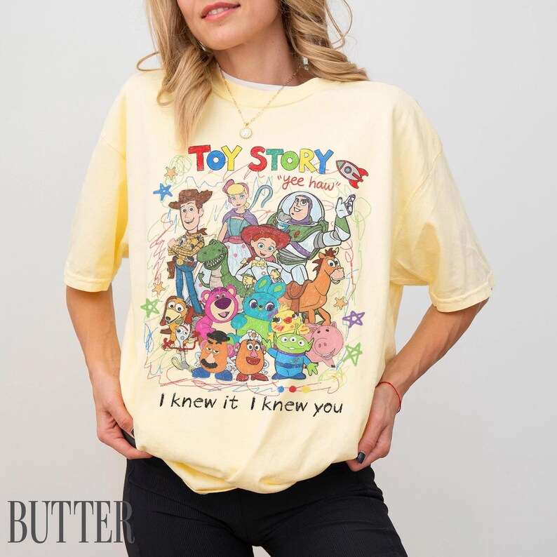 May include: A pale yellow short-sleeve t-shirt featuring a colourful graphic print of Toy Story characters including Woody, Buzz Lightyear, and Jessie. The design includes the text "TOY STORY", "yee haw", and "I knew it I knew you" with scribbled accents.