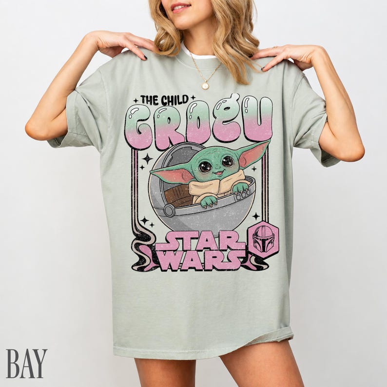 May include: A light sage green t-shirt featuring a graphic print of the character Grogu from Star Wars sitting in a floating pod. The text reads THE CHILD GROGU and STAR WARS in a stylized pink and green font, with a small helmet icon included.