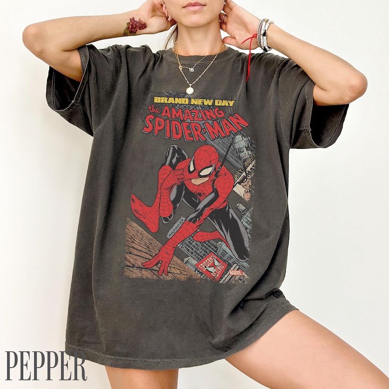 May include: Dark gray t-shirt featuring a comic book graphic of Spider-Man. The design includes the text "BRAND NEW DAY the AMAZING SPIDER-MAN" in red and yellow. The shirt has a vintage, worn look.