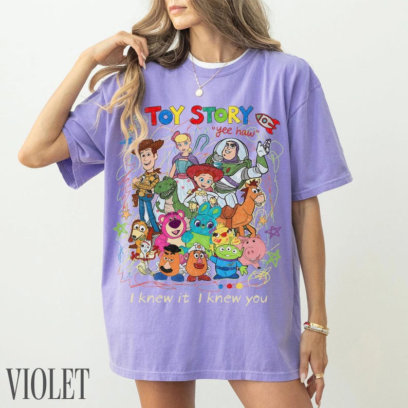 May include: A light purple short-sleeve t-shirt featuring a colourful graphic print of Toy Story characters including Woody, Buzz Lightyear, and Jessie. The text reads Toy Story, yee haw, and I knew it I knew you with scribbled star accents.