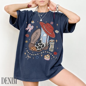 May include: A dark blue short-sleeve t-shirt featuring a graphic print of a cow-print cowboy boot, a red hat, blocks with the letters T, S, and the number 13, and the text TAYLOR. The design includes small stars, bows, and a beaded string.
