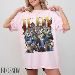 May include: Light pink t-shirt featuring a graphic of Judy Hopps from Zootopia. The design includes the name "JUDY" in gold lettering and several cartoon images of Judy Hopps. The word "Blossom" is printed at the bottom.