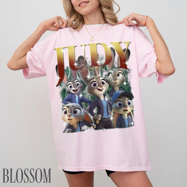May include: Light pink t-shirt featuring a graphic of Judy Hopps from Zootopia. The design includes the name "JUDY" in gold lettering and several cartoon images of Judy Hopps. The word "Blossom" is printed at the bottom.