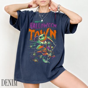 K&ouml;nnte beinhalten: Ein dunkelblaues Kurzarm-T-Shirt mit einem Grafikdruck und dem Text "NIGHT IN HALLOWEEN TOWN" in Lila und Orange. Das Design zeigt Illustrationen von Charakteren wie Jack Skellington, Sally und Oogie Boogie in leuchtenden Farben.