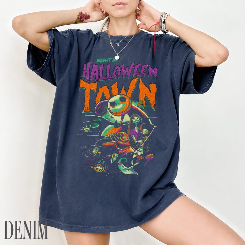 K&ouml;nnte beinhalten: Ein dunkelblaues Kurzarm-T-Shirt mit einem Grafikdruck und dem Text "NIGHT IN HALLOWEEN TOWN" in Lila und Orange. Das Design zeigt Illustrationen von Charakteren wie Jack Skellington, Sally und Oogie Boogie in leuchtenden Farben.