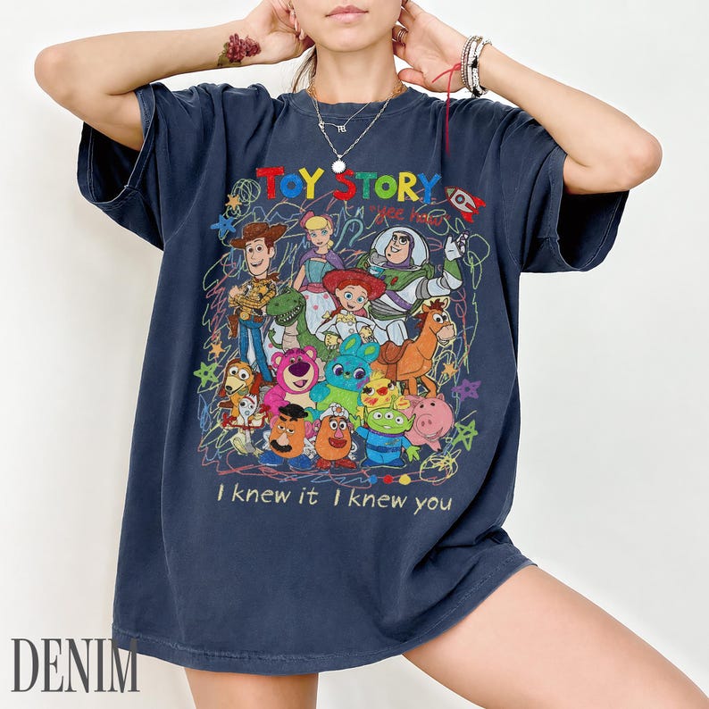May include: A dark blue short-sleeve t-shirt featuring a colourful graphic print of Toy Story characters including Woody, Buzz Lightyear, and Jessie. The design includes scribbled line art, stars, and the text "TOY STORY" and "I knew it I knew you".