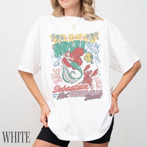 Puede incluir: Una camiseta blanca de manga corta con un estampado gr&aacute;fico colorido de Ariel, Flounder y Sebasti&aacute;n. El texto dice Atlantica Ocean Tour 1991, Sebastian and the Hot Crustacean Band. El dise&ntilde;o incluye ilustraciones de corales y burbujas de estilo vintage.