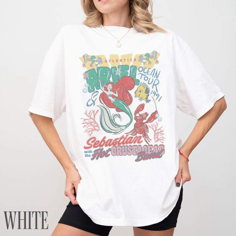 Puede incluir: Una camiseta blanca de manga corta con un estampado gr&aacute;fico colorido de Ariel, Flounder y Sebasti&aacute;n. El texto dice Atlantica Ocean Tour 1991, Sebastian and the Hot Crustacean Band. El dise&ntilde;o incluye ilustraciones de corales y burbujas de estilo vintage.