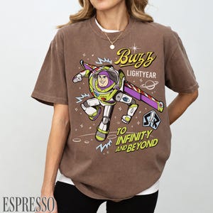 Pode incluir: Camiseta marrom com um desenho do Buzz Lightyear. O design inclui o texto "Buzz Lightyear" e "To Infinity and Beyond". A camiseta tem um visual vintage e desgastado.