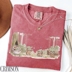Pu&ograve; includere: T-shirt color cremisi con grafica di Grogu. Il design mostra Grogu in un ambiente forestale con altri personaggi. Una collana d'argento con un ciondolo &egrave; drappeggiata sulla maglietta. La maglietta ha un'etichetta Comfort Colors.