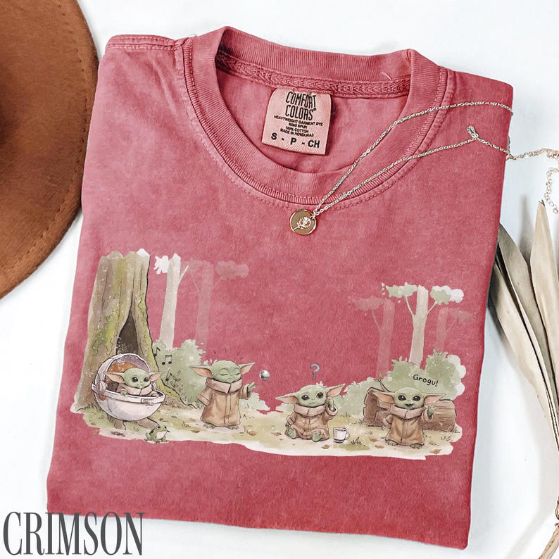 Pu&ograve; includere: T-shirt color cremisi con grafica di Grogu. Il design mostra Grogu in un ambiente forestale con altri personaggi. Una collana d'argento con un ciondolo &egrave; drappeggiata sulla maglietta. La maglietta ha un'etichetta Comfort Colors.