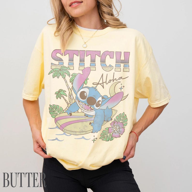 May include: Yellow t-shirt featuring a graphic of Stitch surfing with the word "STITCH" above and "Aloha" below. The design includes palm trees, a crescent moon, and a flower. The shirt is a light yellow color.
