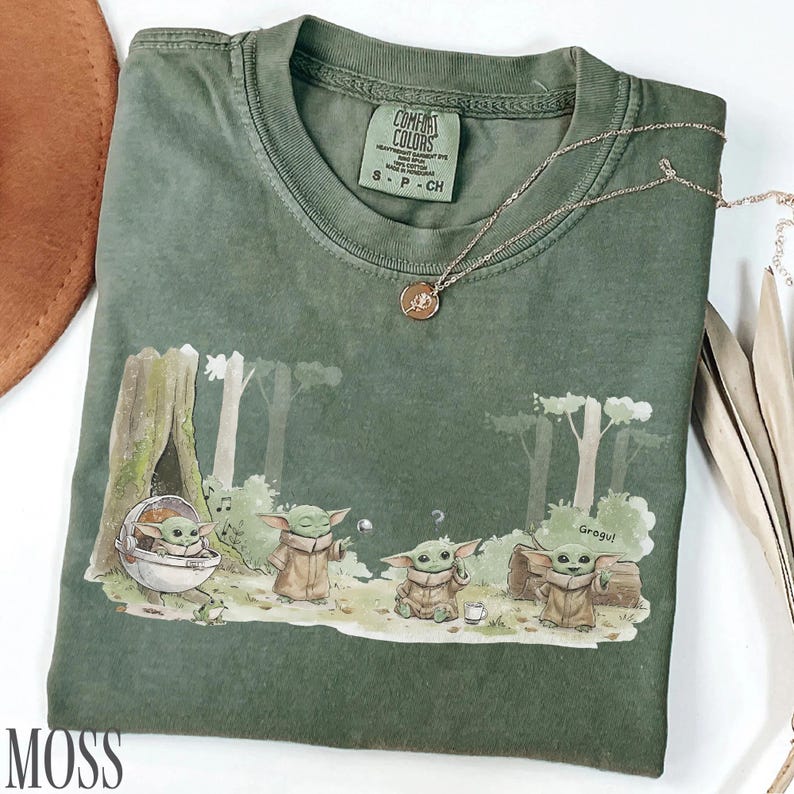 Pu&ograve; includere: T-shirt verde con una grafica di Grogu da The Mandalorian in una scena di foresta. La maglietta ha un girocollo e maniche corte. Una collana d'oro con un ciondolo &egrave; sulla maglietta. La parola "MOSS" &egrave; in basso a sinistra.