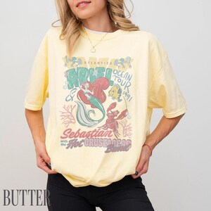 Puede incluir: Una camiseta de manga corta de color amarillo p&aacute;lido con un estampado gr&aacute;fico que incluye el texto Atlantica Ocean Tour 1991, Ariel, Sebastian y the Hot Crustacean Band. El dise&ntilde;o muestra ilustraciones de una sirena, un cangrejo y un pez amarillo.