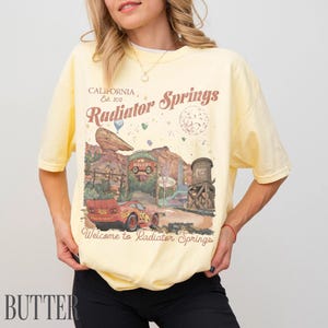 May include: A pale yellow short-sleeve t-shirt featuring a graphic print of Radiator Springs from the movie Cars. The design includes a red race car, desert rock formations, a water tower, and the text California Est. 2012 Radiator Springs.