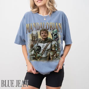 May include: A pale blue t-shirt with a graphic of characters from The Mandalorian. The word "MANDALORIAN" is printed across the top in gold lettering. The shirt also has the words "BLUE JEAN" printed at the bottom.