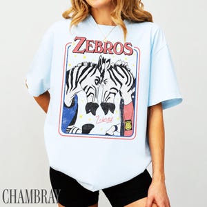Op de afbeelding: Lichtblauw T-shirt met een afbeelding van twee zebra's in een rood en blauw kader. Het woord "ZEBROS" staat in roze boven de zebra's, en het woord "Zebras" staat eronder. Het shirt heeft een relaxte pasvorm.