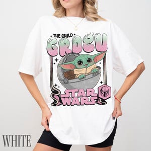 May include: A white short-sleeve t-shirt featuring a graphic print of Grogu from Star Wars inside a floating pod. The text reads THE CHILD GROGU and STAR WARS in a pink and green gradient font, with a small pink Mandalorian helmet icon included.