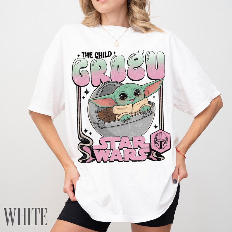 May include: A white short-sleeve t-shirt featuring a graphic print of Grogu from Star Wars inside a floating pod. The text reads THE CHILD GROGU and STAR WARS in a pink and green gradient font, with a small pink Mandalorian helmet icon included.