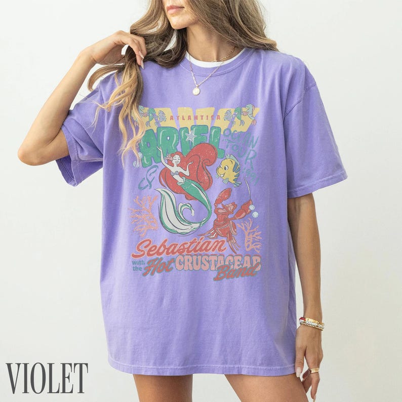 Puede incluir: Una camiseta de manga corta de color violeta claro con un estampado gr&aacute;fico de Ariel, Flounder y Sebasti&aacute;n. El texto dice Atlantica, Ariel, Sebastian with the Hot Crustacean Band. El dise&ntilde;o incluye corales y burbujas en estilo desgastado.