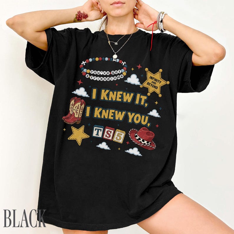 May include: A black t-shirt featuring a graphic print with the text "I KNEW IT, I KNEW YOU," "YOU'VE GOT A FRIEND IN ME," "INFINITY AND BEYOND," and "TS5." The design includes a cowboy boot, a sheriff star, a cowboy hat, and colorful blocks.