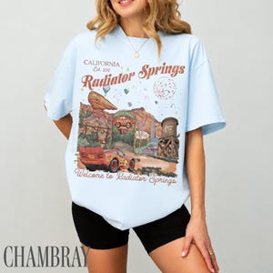 May include: A light blue t-shirt featuring a graphic print of a red race car in a desert landscape. The text reads California Est. 2012, Radiator Springs, and Welcome to Radiator Springs. The design includes a rock formation and a water tower.
