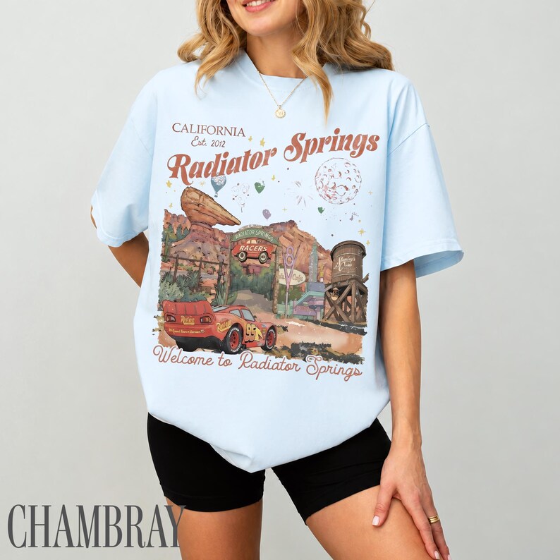 May include: A light blue t-shirt featuring a graphic print of a red race car in a desert landscape. The text reads California Est. 2012, Radiator Springs, and Welcome to Radiator Springs. The design includes a rock formation and a water tower.