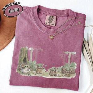 Pu&ograve; includere: T-shirt Comfort Colors color bacca con grafica di Grogu in una scena di foresta. La grafica include Grogu in una capsula e altri personaggi di Grogu. La maglietta ha girocollo e maniche corte. Una collana d'oro &egrave; drappeggiata sulla maglietta.