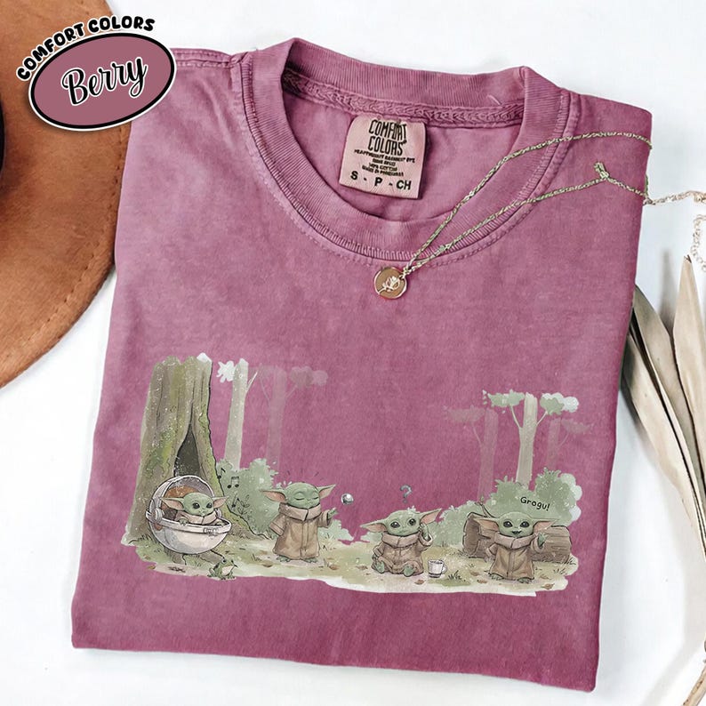 Pu&ograve; includere: T-shirt Comfort Colors color bacca con grafica di Grogu in una scena di foresta. La grafica include Grogu in una capsula e altri personaggi di Grogu. La maglietta ha girocollo e maniche corte. Una collana d'oro &egrave; drappeggiata sulla maglietta.
