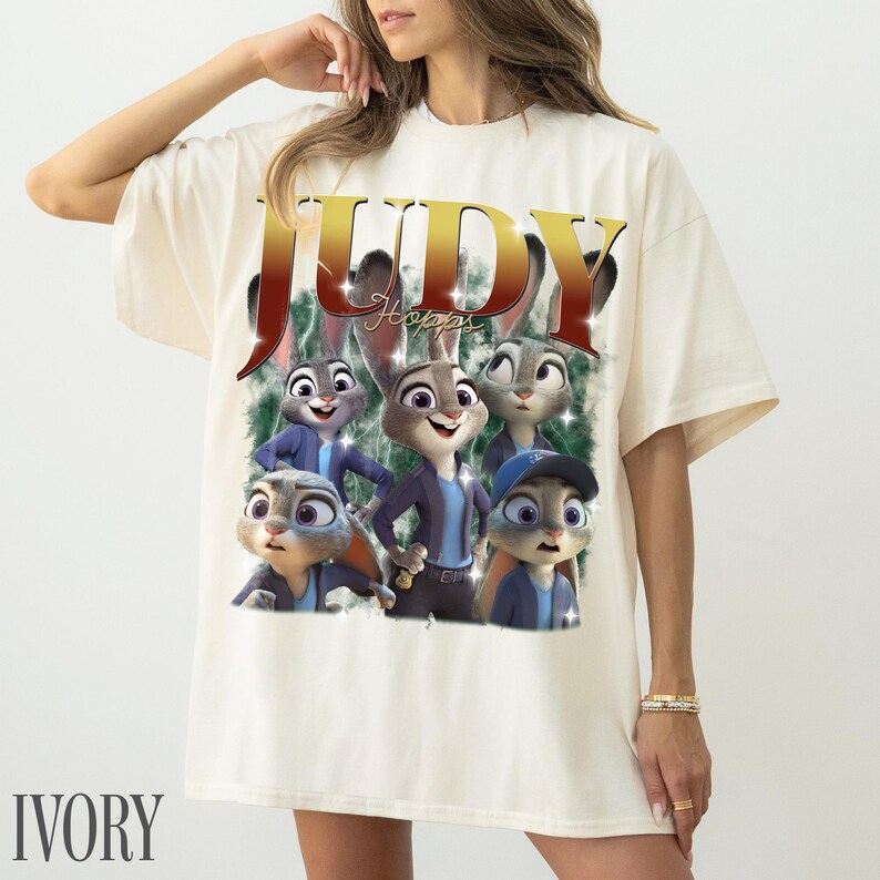 May include: Ivory-colored t-shirt featuring a graphic of Judy Hopps from Zootopia. The design includes the word "JUDY" in large, gold letters and images of Judy Hopps and other characters. The word "IVORY" is printed at the bottom.
