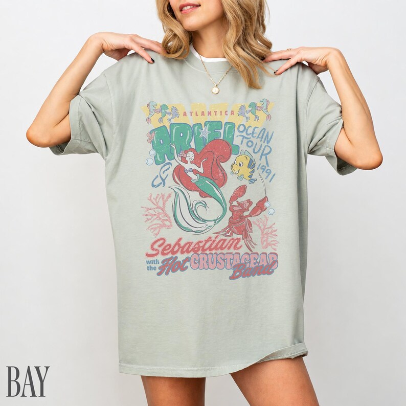 Puede incluir: Una camiseta de manga corta color verde salvia con un estampado gr&aacute;fico de Ariel, Flounder y Sebasti&aacute;n. El texto dice Atlantica Ocean Tour 1991, Sebastian with the Hot Crustacean Band. El dise&ntilde;o incluye ilustraciones de coral y burbujas en rojo y azul.