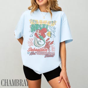 Puede incluir: Una camiseta de manga corta azul claro con un estampado gr&aacute;fico de Ariel, Flounder y Sebasti&aacute;n. El texto dice "Ariel Ocean Tour 1991" y "Sebastian with the Hot Crustacean Band". El dise&ntilde;o incluye ilustraciones de coral y burbujas.